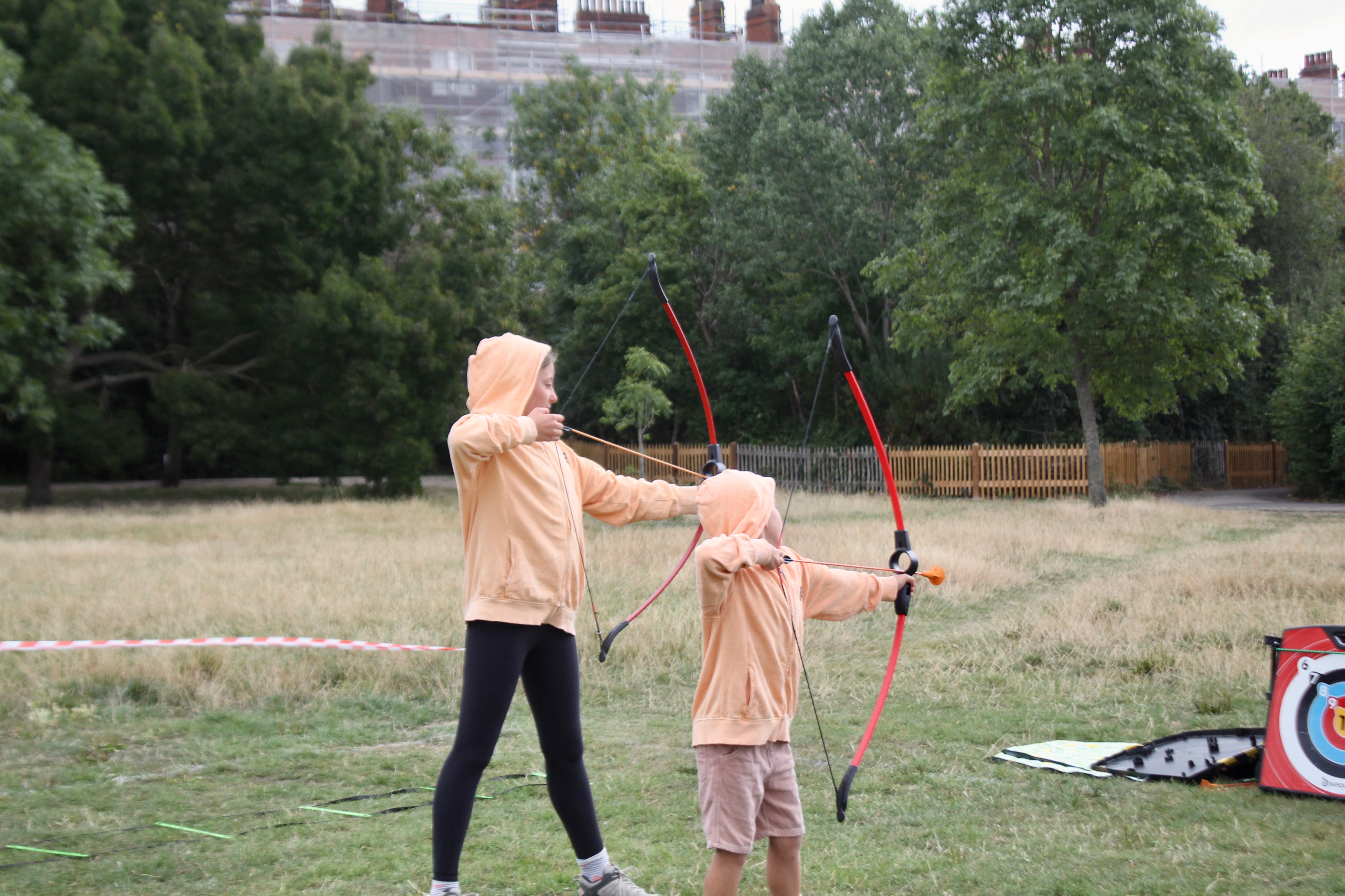 Photo of a young girl and a little boy wearing an orange jumper, drawing a bow toy and aiming at a target in a public park