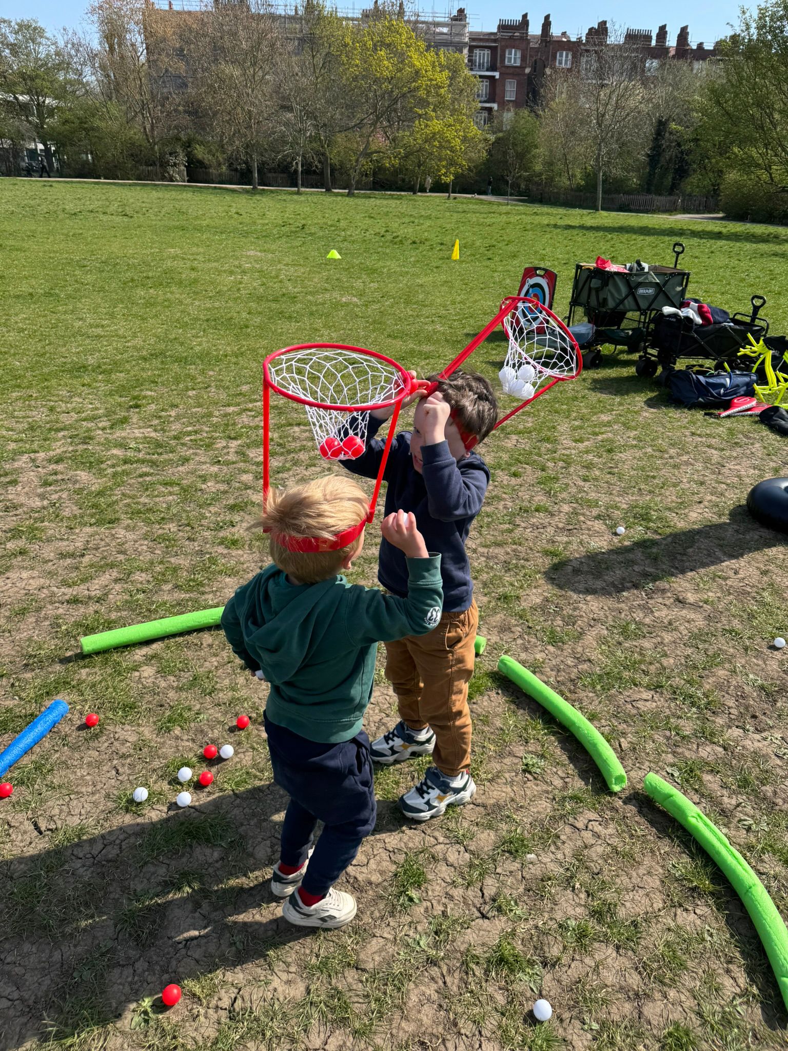 Photo of two children in a park on a sunny day, each wearing a mesh basket on their head and playing a game where they have to pick up red and white balls placed on the ground in a marked area and put them in the other's basket